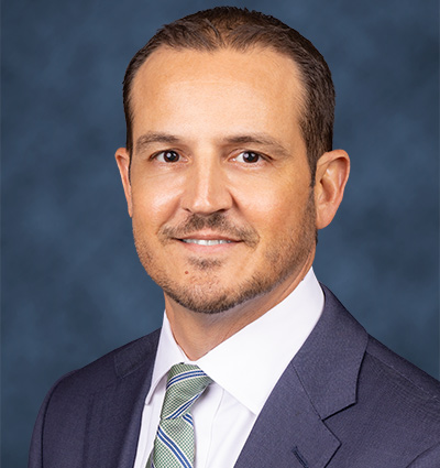 A man smiling at the camera, he is wearing a blue jacket, white shirt, and green and blue tie