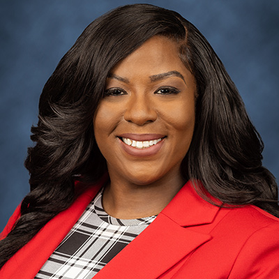 A woman smiling at the camera, she has long black hair, a bright jacket, and a checkered shirt