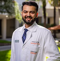 A headshot of a male physician, he smiles at the camera. He is wearing a white coat and has short dark hair