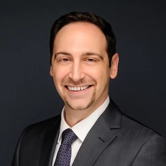 A headshot of a professional man, he has on a dark suit coat, white shirt, and dark tie, he smiles at the camera