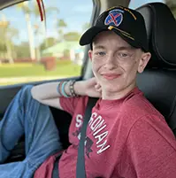A young boy sitting in a car, he smiles at the camera, he has on jeans, a red shirt, and a hat