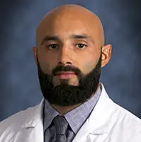 A headshot of a medical professional, he is looking at the camera, has on a white coat, dress shirt, and tie