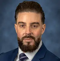 A headshot of a man, he has dark hair, facial hair, a suit jacket, dress shirt, tie