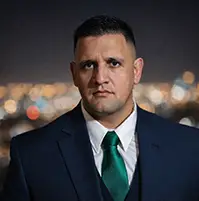 A man looking at the camera, he has on a suit with a green tie, he has short dark hair