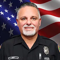 A headshot of a police officer, there's is an American flag behind him, he smiles at the camera