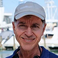 A closeup of a man, he's standing in front of boats, he has on a gray hat and smiles at the camera