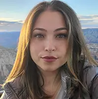 A woman looking at the camera, there is a mountain landscape behind her, she has long light brown hair