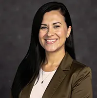 A woman smiling at the camera, she has long dark hair, she wears an olive colored jacket and white top