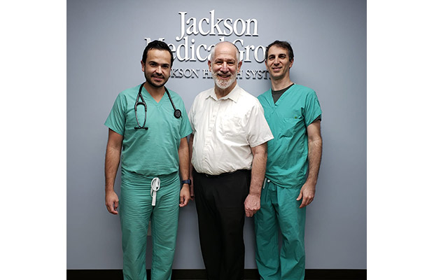 Two physicians standing next to an older male patient, they stand in front of a sign that reads Jackson Medical Group