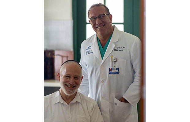 A physician smiling and standing behind an older male who is smiling at the camera