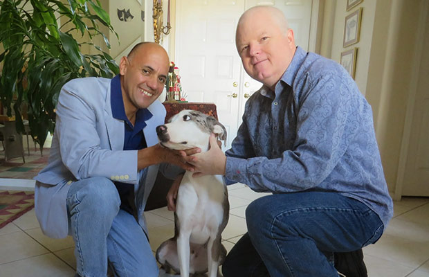 Two men kneeling and petting a dog, they both smile at the camera
