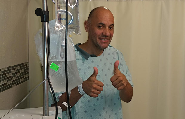 A men wearing a hospital gown, holding two thumbs up and smiling at the camera