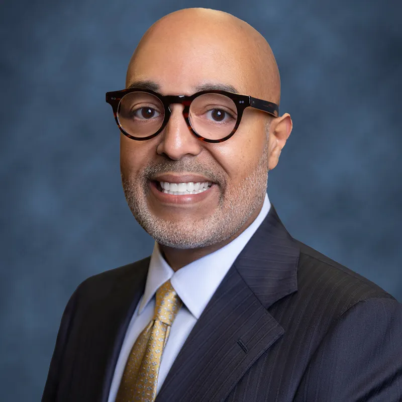 Professional headshot of Zambrana smiling, wearing round glasses, a dark pinstripe suit, and a gold patterned tie, against a blue-gray studio background