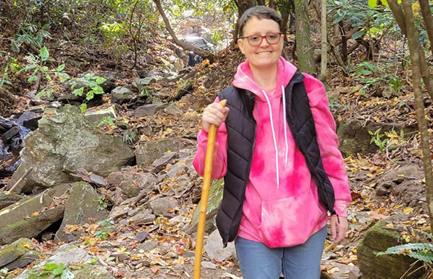 A person hiking outdoors, they smile at the camera and are holing onto a wooden walking stick