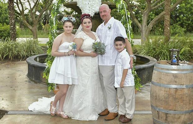 A man, woman, a young girl, and young boy, standing next to each other in wedding attire smiling at the camera; they are outdoors