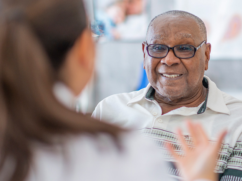 An older male smiling as he listens to his physician, you see the back of his physicians head