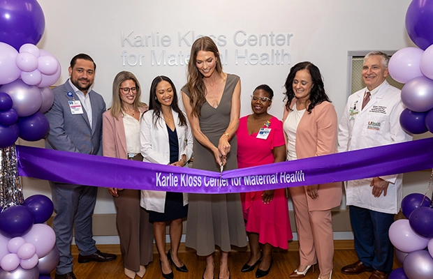 Group of healthcare professionals and a woman standing together at an indoor ribbon-cutting ceremony, surrounded by purple and white balloon decorations.