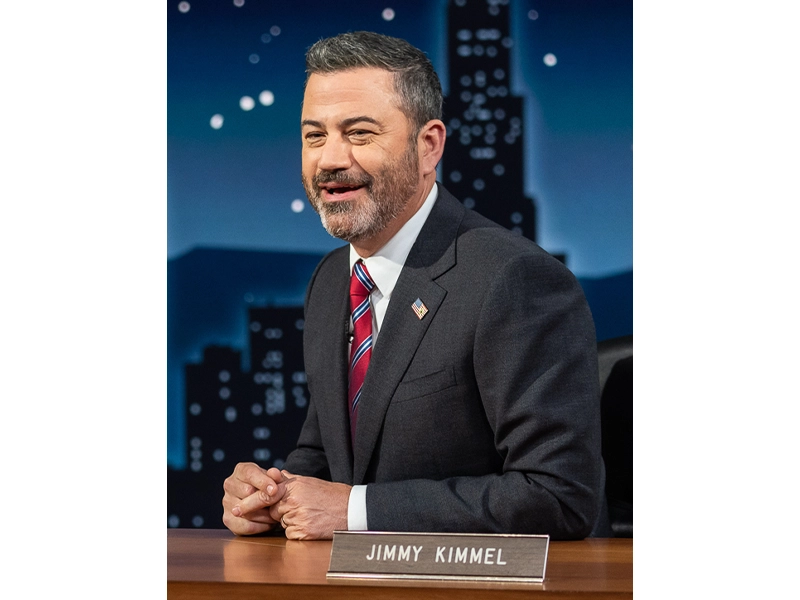 Photograph of Jimmy Kimmel during a show, he is seen sitting at a desk, wearing a black suit with a red tie