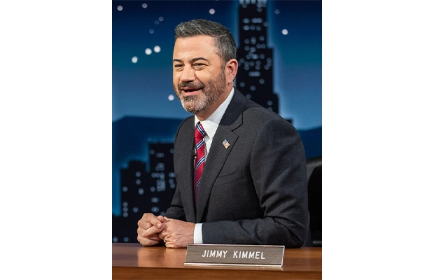 Photograph of Jimmy Kimmel during a show, he is seen sitting at a desk, wearing a black suit with a red tie