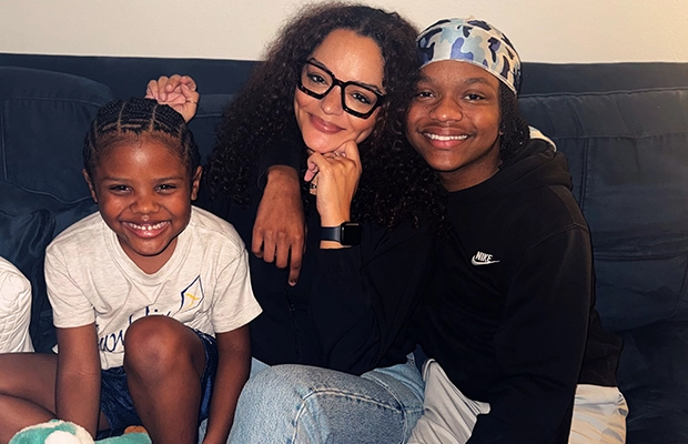 A young boy posing next to a woman and a girl who are seated on a sofa.