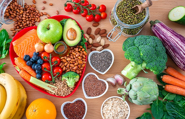 Overhead view of a group of food on a wooden table, there are numerous foods in a heart shaped plate. The food items include extra virgin olive oil, legumes, avocado, berries, nuts and seeds, salmon, broccoli, carrot, and more.