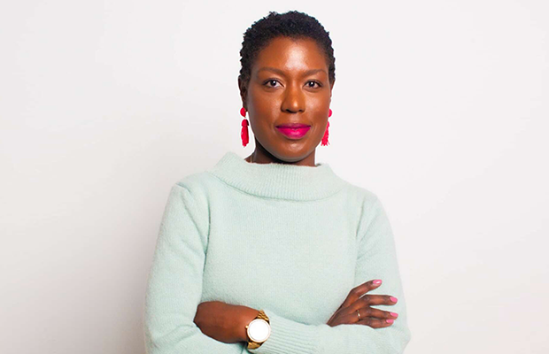 A woman looking at the camera, her arms are crossed, she wears a long light green top, she has on pink earrings