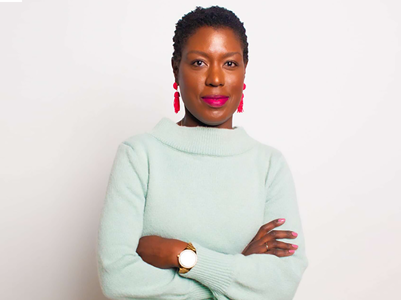 A woman looking at the camera, her arms are crossed, she wears a long light green top, she has on pink earrings