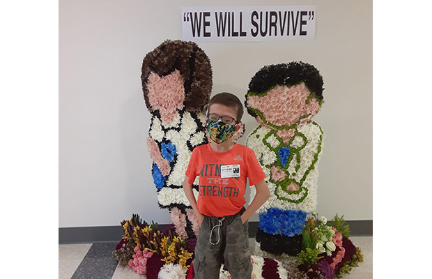 A young boy standing between two physician statues made of flowers, behind there is a sign that reads we will survive