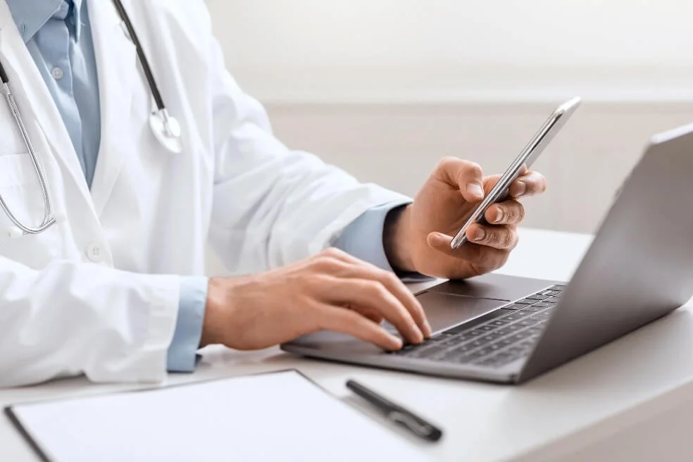 A closeup of a medical professional using a laptop and mobile phone