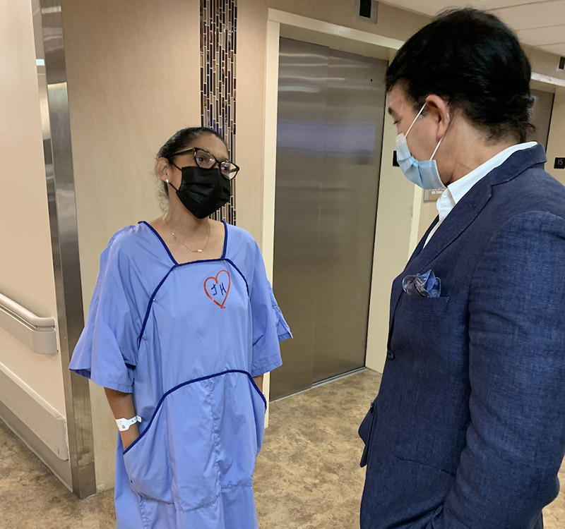 A patient in a hospital wearing a long blue gown, she has on glasses and a face mask, she is speaking with a man who has on a face covering and wearing a dark blue suit jacket
