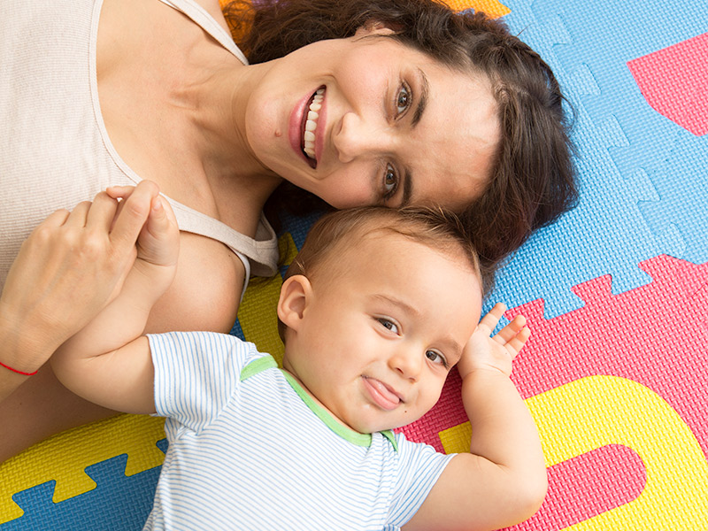 Maria and her baby smiling at the camera as they lay down on a mat and hold hands