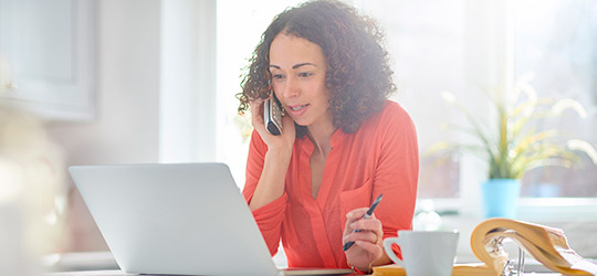 Woman in her kitchen holding a phone within her right hand, she looks at her laptop and is holding a pen in her left hand
