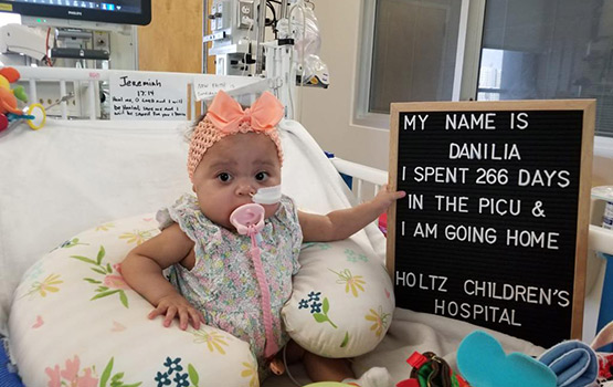 A baby sitting on a medical bed, she holds a sign that reads My name is Danilia, I spent 266 days in the PICU and I am going home, Holtz Children's Hospital