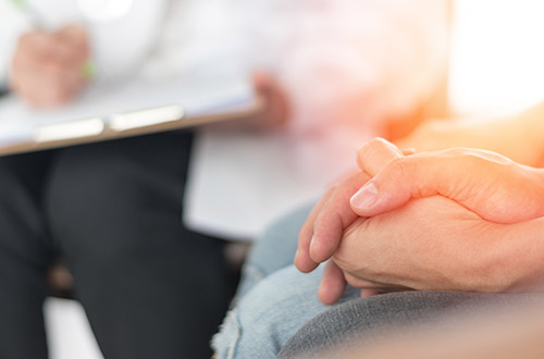 A closeup of two hands intertwined as you see a doctor writing in their chart in the background