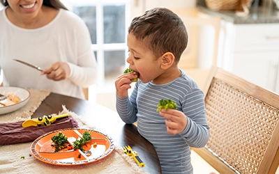 Mom smiling as she watches her son eat vegetables.