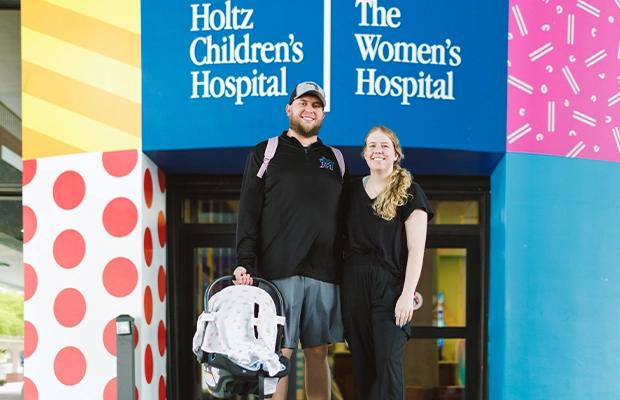 A couple posing for the camera in front of the hospital while holding their baby.