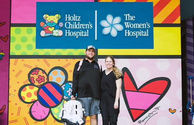A couple posing for the camera in front of the hospital while holding their baby.