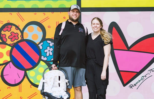 A couple posing for the camera in front of the hospital while holding their baby.
