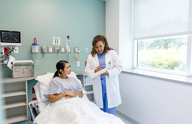 A patient laying on a hospital bed, there's a medical professional speaking with her and showing her something on a clipboard