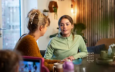A young girl sitting at a dining room table speaking to a woman, they look to be at home