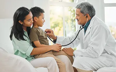 A medical professional using a stethoscope to check a young boys heart, the boys mom has him on her lap