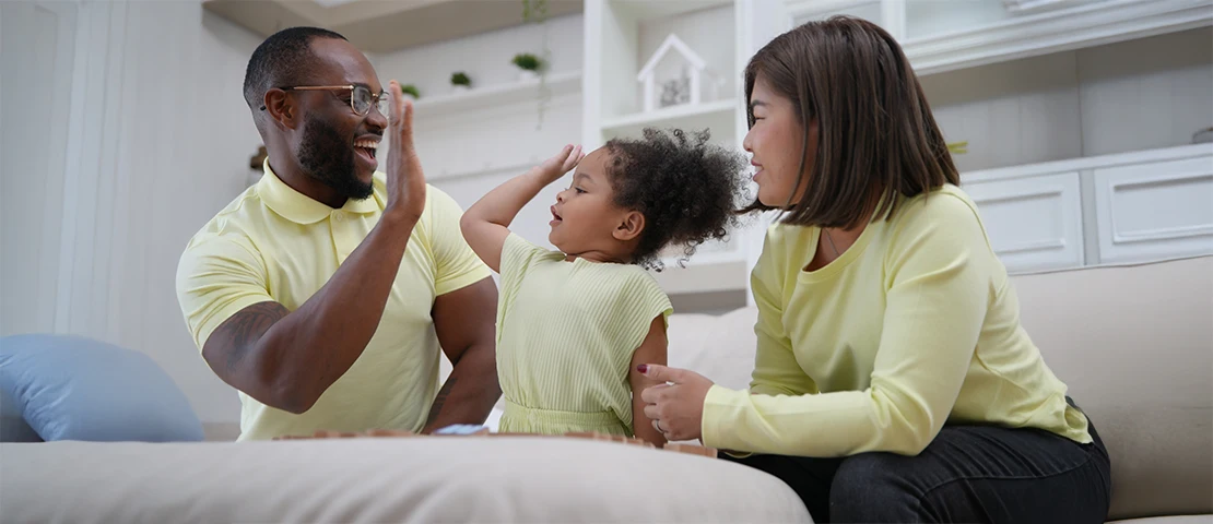 A parent giving his young child a high five, the mom sits next to the child