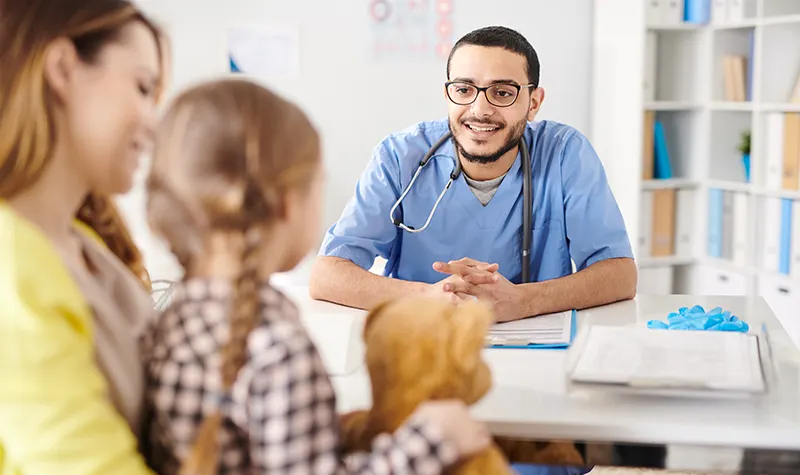 A smiling male healthcare provider in blue scrubs with a stethoscope consulting with a mother and young girl holding a stuffed animal in a pediatric clinic.