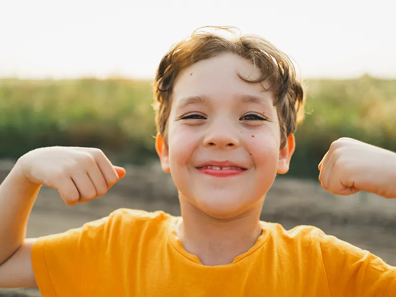 A cheerful young boy in an orange t-shirt flexing both arms outdoors, smiling confidently at the camera.