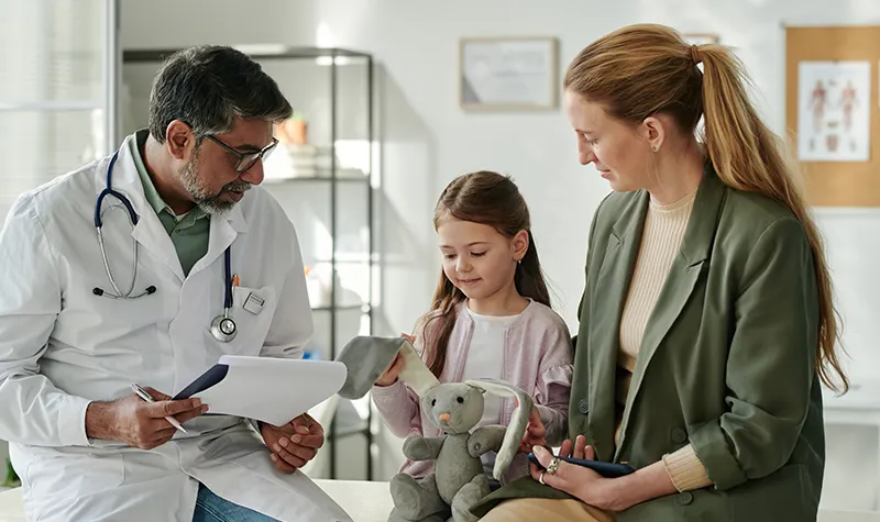 A physician speaking to a mom and young girl at a physicians office, the young girl is holding a stuffed bunny