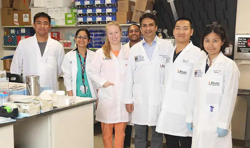 A group of medical researchers wearing lab coats stand together in a laboratory, smiling for a group photo. Lab equipment, shelves with supplies, and research materials are visible in the background