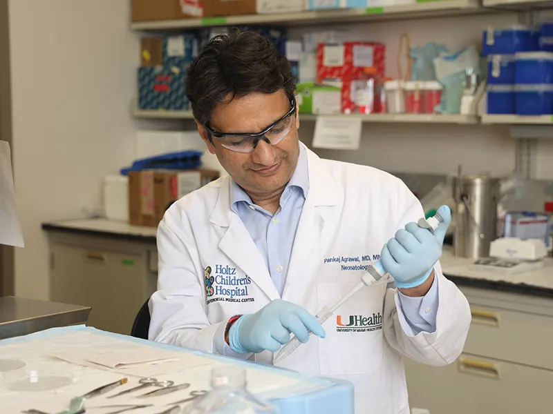 A medical researcher in a lab coat and safety glasses uses a pipette while working at a laboratory bench. Lab equipment and organized supplies are visible behind him