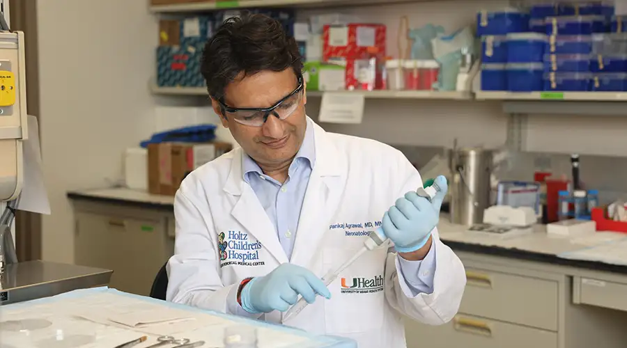 A medical researcher in a lab coat and safety glasses uses a pipette while working at a laboratory bench. Lab equipment and organized supplies are visible behind him