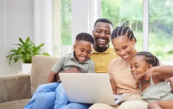 A smiling family of four, they are sitting on a couch at home and looking at a laptop screen