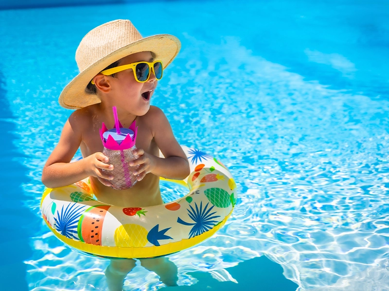 A boy in the pool wearing a hat, goggles, float, holding a drink